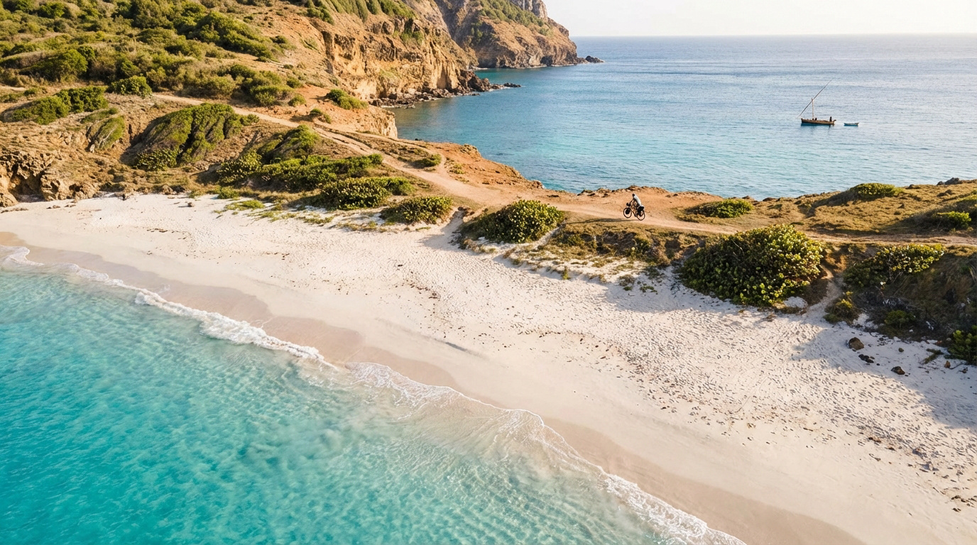 Vue aérienne d'une plage de sable blanc et d'eaux turquoise à La Désirade. Un cycliste roule sur un sentier côtier. Un voilier est en mer.